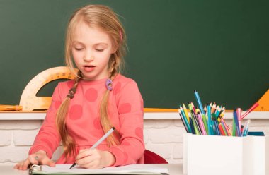 İlkokuldan bir çocuk ilkokulda masada oturuyor, not defterine yazıyor ya da çiziyor. Okulda tahtaya yakın bir okul çocuğu.