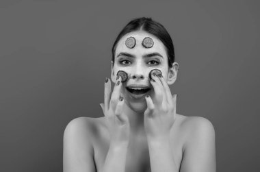 Close up beauty portrait of a beautiful woman applying face cream and facial mask of cucumber. Mask for skin lifting and anti-aging effect, studio background. Anti aging cream. Facial treatment