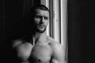 Oturma odasında pencere perdelerinde seksi çıplak genç adam. Çıplak erkek vücudu. Seksi manken. Seksi vücutlu genç kaslı adam. Çıplak göğüs. Kaslı erkek gövdesi, çıplak omuzlar.