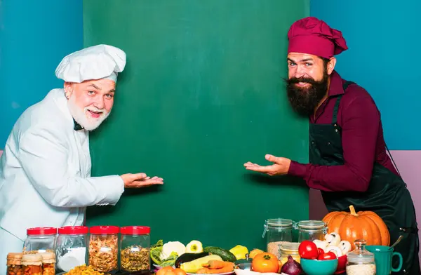 Stock image Two chefs in cook hat pointing spoon on blank board menu. Kitchen chef