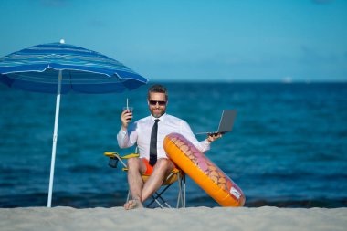 Yaz tatillerinde başarılı bir iş adamı. Komik iş adamı deniz sahilinde yaz kokteyli içip dizüstü bilgisayar kullanıyor. Yaz işi konsepti. Sıcak yaz. Çılgınca. Uzak çevrimiçi çalışma