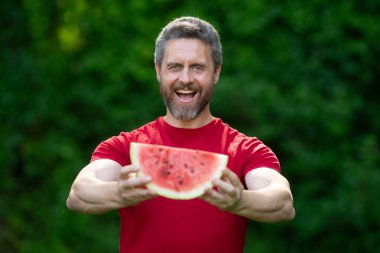 Karpuzlu komik erkek suratı, yaklaş. Yazlık parkta karpuz yiyen heyecanlı bir adam. Karpuz yiyen bir adam. 40 'ların adamı dışarıda meyve yiyor. Karpuz seven bir adam