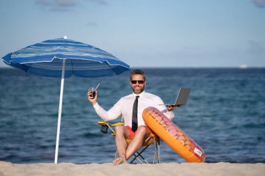 Yaz tatillerinde başarılı bir iş adamı. Komik iş adamı deniz sahilinde yaz kokteyli içip dizüstü bilgisayar kullanıyor. Yaz işi konsepti. Sıcak yaz. Çılgınca. Uzak çevrimiçi çalışma