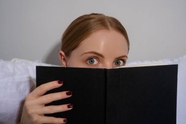 Açık bir kitabın arkasına saklanan güzel bir kızın portresi. Yüzünü kitapla örten kadın. Kitabın arkasına saklanan kız. Kitaplı güzel bir genç kadının yakın plan portresi.