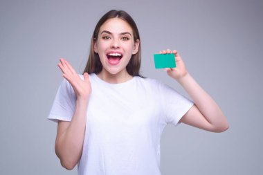 Yüzünün yanında kredi kartı tutan beyaz tişörtlü genç bir kadın. Gri arka planda izole edilmiş. Zeka ya da banka kartını öde. Banka kartı. Plastik kredi kartı