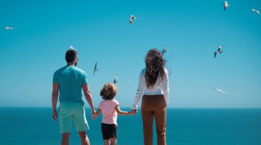 Gökyüzündeki plajda aile manzarası. İnsanlar yaz tatilinde eğleniyor. Baba, anne ve çocuk mavi denize karşı el ele tutuşuyorlar. Tatil seyahati kavramı