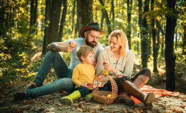 Küçük çocuklu genç bir aile sonbahar doğasında gün batımında piknik yapıyor. Çocuk için içinde yemek ve oyuncaklar olan bir sepet var. Gülümseyen genç bir aile sonbahar günü piknik yapıyor.
