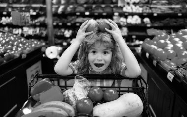 Komik çocuk dükkanda elma seçiyor. Komik küçük çocuk gülüşü ve elma tutuşu. Çocuk süpermarkette alışveriş yapıyor. Arabalı küçük çocuk marketten taze sebze seçiyor.