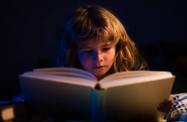 Uyumadan önce yatakta kitap okuyan bir çocuğun yakın plan portresi. Evdeki küçük çocuk yatakta uzanıp kitap okuyor.