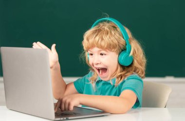 Heyecanlı kulaklıklı çocuk, masada oturur, okulda dizüstü bilgisayarda çalışır, klever şaşkın çocuk internet derslerini kullanarak not defterine kulaklık takar.