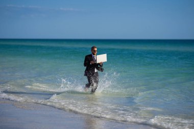 Deniz suyunda dizüstü bilgisayarı olan takım elbiseli bir iş adamı. Seyahat turizmi ve iş konsepti. Denizde dizüstü bilgisayarı olan çılgın bir ofis çalışanı. Yaz tatilindeki çılgın iş adamı.