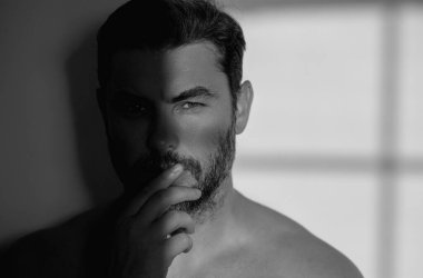 Yüz kreminden sonra tatmin olmuş genç bir adamın yakından güzel portresi. Çekici esmer adam tenine dokunuyor. Sakallı ve modern saç kesimli erkek modeller. Erkek kozmetik ve güzellik konsepti
