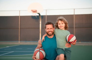Baba oğul basketbol oynuyor. Babamın portresi, elinde basketbol topu tutan küçük çocuğu kucaklıyor.