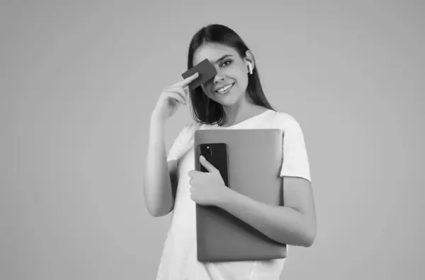 stock image Young woman in white t-shirt holding credit card near face and laptop, isolated over gray background. Pay wit credit or debit card. Bank card. Plastic credit card