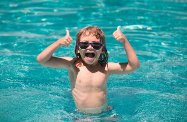 Yaz yüzme havuzunda bir çocuk. Başparmakları havada, yüzme havuzunda yüzen tatlı çocuk, fotokopi alanı olan yaz suyu arka planı. Komik çocuk suratı. Başparmaklar yukarı.