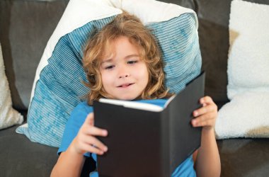 Hayali çocuk. Çocuk oturma odasında kitap okuyor. Çocuklar kitap okur. Küçük çocuk güneşli oturma odasında kanepede oturmuş masal kitabındaki resimleri izliyor.