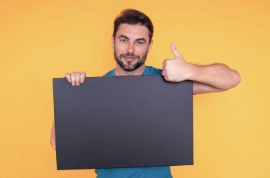 Başparmağı havada bir adam stüdyo sarı arka planda pankart tutuyor. Elinde boş bir poster tutan adamın portresi. Boş poster gösteren, işaret parmağı sallayan bir adam. Pano, reklam panosu ya da afiş sunan adam