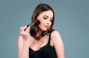 Fashion portrait of beautiful glamor model with makeup blusher brush and stylish makeup on the face isolated on gray background. Girl hold blush blusher, apply powder