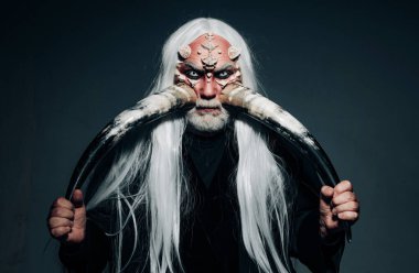 İki kırmızı boynuz tutan yaşlı bir adam. Şeytan kafası siyaha izole edilmiş. Eski simya. Şeytan boynuzları. Cadılar Bayramı canavarı gibi giyinmiş yaşlı sakallı bir adam. Siyah zemin üzerinde Druid