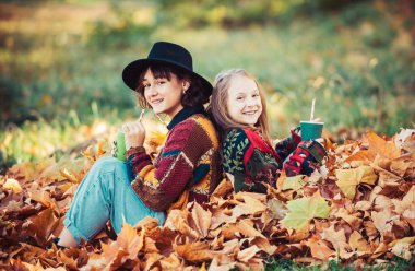 Sonbahar sarı yapraklı mutlu kardeşler. Düşmüş sarı yapraklar. Elinde portakal yaprağı olan kız kardeşiyle birlikte gülen genç bir kız ormanda sonbahar yapraklarının üzerinde oturuyor. Kardeşler güz doğasının tadını çıkarıyor.
