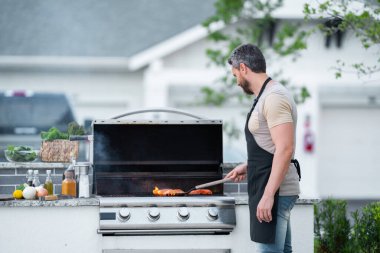 Yakışıklı adamın kırpılmış görüntüsü arka bahçede ızgara yapıyor. Barbekü partisi. Mangal et, piknik için ızgara. Barbeküde kızartılmış. Adam evin bahçesinde barbekü hazırlıyor.