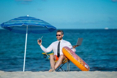 Yaz tatillerinde başarılı bir iş adamı. Komik iş adamı deniz sahilinde yaz kokteyli içip dizüstü bilgisayar kullanıyor. Yaz işi konsepti. Sıcak yaz. Çılgınca. Uzak çevrimiçi çalışma