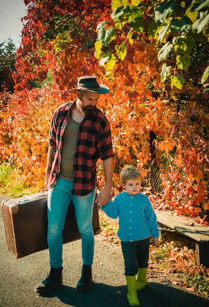 stock image Loving dad carefuly holds hand of his little son. Bearded father wearing hipster outfit and holding large vintage travel bag. Family leisure. Parenting. Parenthood. Autumnal ivy leaves background