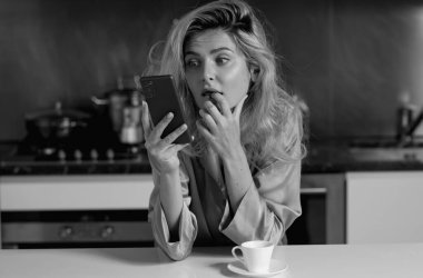 Şaşırmış bir kadın sabah mutfakta oturmuş telefon izliyor ve akıllı telefonlardan sohbet ediyor.