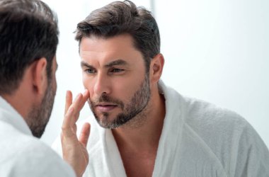 Bin yıllık İspanyol adam aynaya, yüz derisine ve sakalına bakıyor. Erkek güzellik ürünleri. Cilt bakımı, spa. Güzel bir adamın güzel portresi. Spa modeli, nemlendirici krem