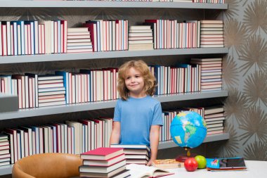 Kütüphanede yığınla kitabı olan okul çocuğu. Okul ve çocuklar. Kitap okuyan sevimli sarışın çocuk. Bilgi günü