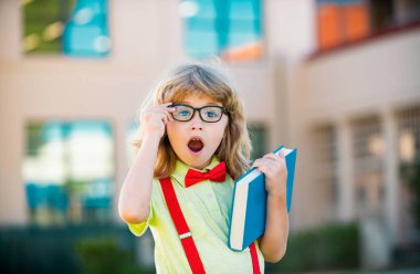 Büyük sırt çantalı ve kitaplı neşeli küçük çocuk okula karşı eğleniyor. Okula dönelim. Eğitim ve okuma kavramı