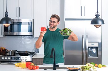 Yakışıklı adam mutfakta salata pişiriyor. Mutfağa sebzeyle yaslanan adam. Mutfakta sebze malzemeleriyle yemek pişiren sıradan bir adamın portresi. Evde salata hazırlayan sıradan bir adam.