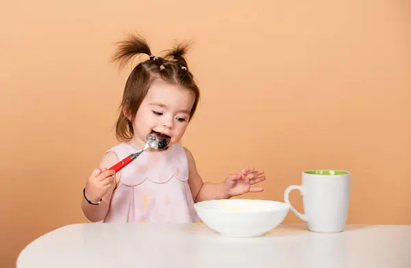 stock image Baby girl with spoon on studio, isolated on beige yellow. Cute funny babies eating, baby food