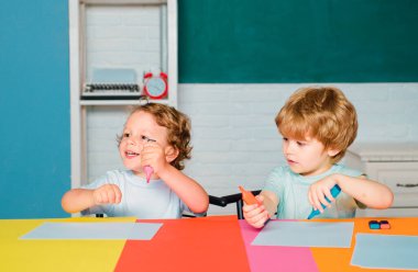 Neşeli, gülümseyen küçük bir oğlan ve küçük tatlı bir kız tahtaya karşı eğleniyor, okul kavramı. Okula ilk gelişim. Okul malzemeleriyle masasının yanında gülen surat ifadesiyle küçük bir çocuk.