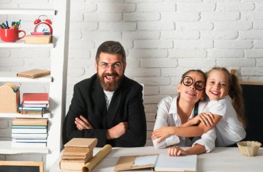 Baba, küçük ilkokul öğrencilerine, kızların okul ödevlerini hazırlamalarına, masada yan yana oturmalarına yardım ediyor. Mutlu küçük çocuklar babalarıyla evde eğitim görüyor.