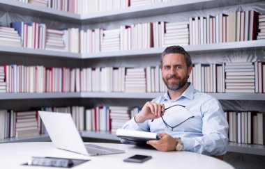 Kütüphanede kitabı olan bir öğretmenin portresi. Üniversite kütüphanesinde yakışıklı bir öğretmen. Öğretmenler Günü. Öğretmen ders veriyor. Kütüphanede öğretmen. Kitaplıktaki üniversite kütüphanesinde özel öğretmen.