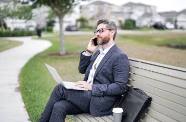 İş konuşmaları. İş adamı telefonda konuşuyor. Parktaki bankta oturuyor. Takım elbiseli bir iş adamı dışarıdaki telefonu arıyor. İş adamı konuşması. Parktaki bankta otur. Takım elbiseli bir iş adamı parkta telefonda konuşuyor.