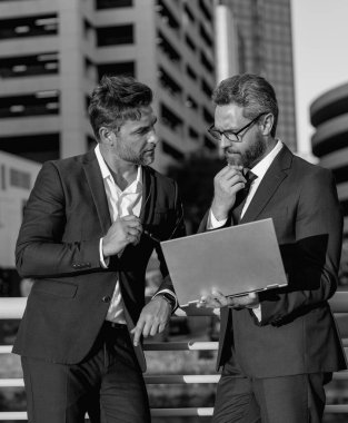 Klasik takım elbiseli iki yakışıklı iş adamı dizüstü bilgisayar kullanıyor. Şehir manzarası yakınında dizüstü bilgisayar kullanan iş adamları. İş adamları toplantıda iletişim kuruyor. İş ortakları toplantısı