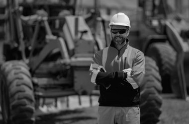 Kazıcının yanında insan üreticisi var. Kazıcılı inşaat işçisi. Miğferli miğfer. Şapkalı bir işçi. İnşaat miğferinde portre mekaniği işçisi. Arkaplanda makine üreticisi