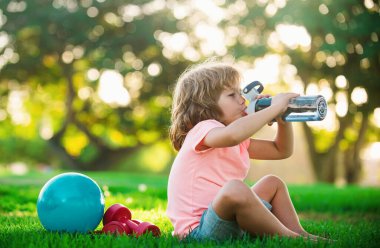 Dambılı ve topları parkta su içen sportif bir çocuk. Çocuklar Spor. Çocuk sağlığı, sağlık ve enerji, halterli egzersizler. Küçük çocuklar için sağlıklı bir yaşam tarzı