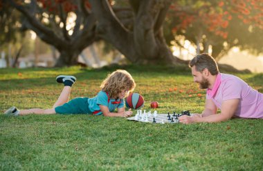 Oğlum çimlere uzanıp babasıyla satranç oynuyor. Çocuk satranç oynamayı öğreniyor. Küçük çocuk satranç oynar ya da düşünür. Şah mat.