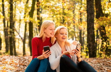İki heyecanlı genç kız sonbahar parkında otururken cep telefonu kullanıyor ve parmakla işaret ediyor.