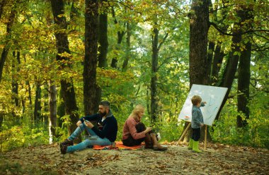 Boyama becerileri. Anne ve baba çocuk resim yaparken parkta çalışıyor. Dinlenme ve hobi konsepti. İş ve mesleki meslek. Küçük oğul doğada resim çiziyor. Sanat ve kendini ifade. Yetenek geliştirme.