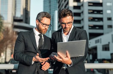 Klasik takım elbiseli iki yakışıklı iş adamı dizüstü bilgisayar kullanıyor. Şehir manzarası yakınında dizüstü bilgisayar kullanan iş adamları. İş adamları toplantıda iletişim kuruyor. İş ortakları toplantısı