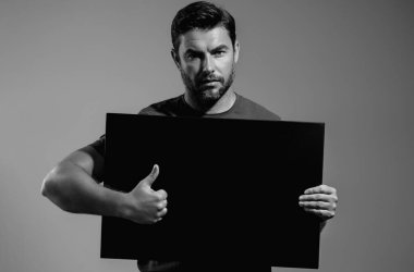 Stüdyonun gri arka planında baş parmağı havada bir adam var. Elinde boş bir poster tutan adamın portresi. Boş poster gösteren, işaret parmağı sallayan bir adam. Pano, reklam panosu ya da afiş sunan adam