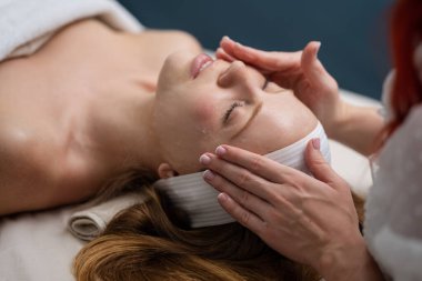 Yüz kilinden maske takan spa kadını, yakın plan. Güzellik kliniğinde cilt masajı. SPA salonunda yüz gerdirme. Kozmetik uzmanı bir kız yüz masajı yapıyor. Kozmetik prosedürü. Güzellik kozmetolojisi