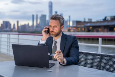 Bilgisayarda çalışan ve dışarıda telefonla konuşan bir iş adamı. İş konuşmaları. Meşgul iş adamı telefonda konuşuyor. Adam iş e-postası ve sörf yapmak için akıllı telefon kullanıyor.