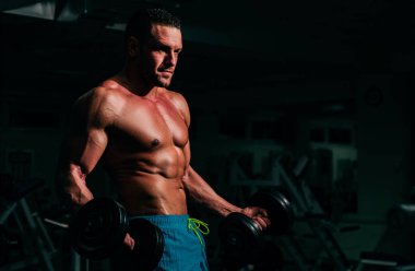 Spor salonunda dambılı olan güçlü bir adam. Vücut geliştirici pazıları egzersizlerle çalıştırıyor. Atletik vücutlu bir sporcu.