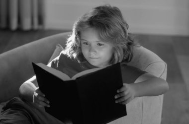 Interesting kids book story. Cute kid in casual clothes reading a book while lying on a sofa in the room. Concept of education and reading