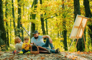 Aile pikniği. Anne, baba ve oğul yeşil parkta resim çiziyor. Aile günü, doğadaki aile aktiviteleri. Atıştırma zamanı. Öğle yemeği için elmalar. Eğitim açık havada